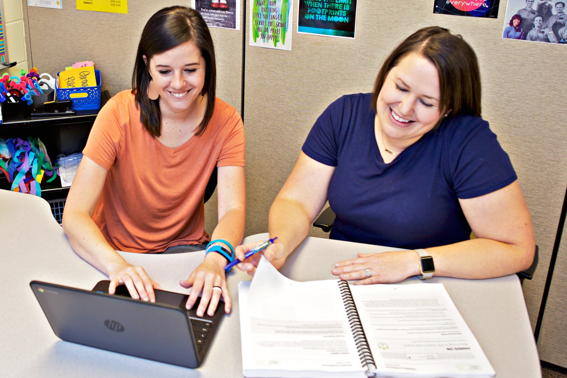 Two women in an office, one using a laptop and another with a notebook, smiling and discussing tutoring services.
