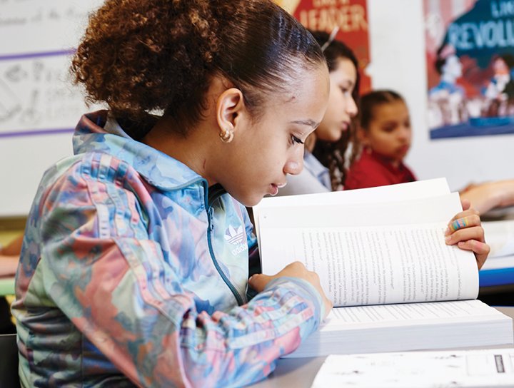 A young student with curly hair reads a book intently in a middle school literacy classroom, with peers in the background.