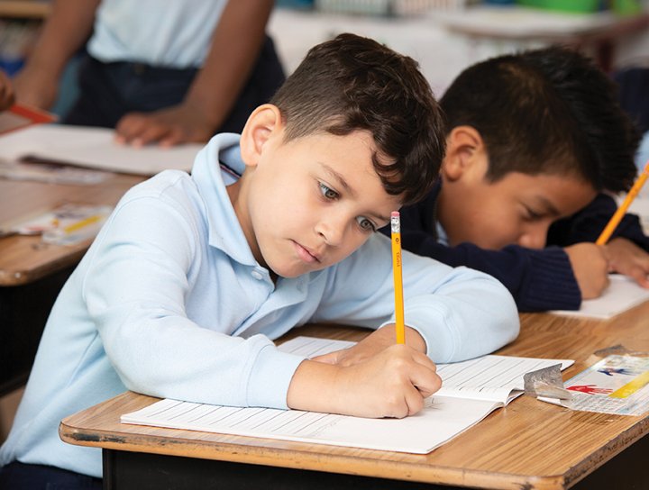 A young boy in a blue shirt writes in a notebook with a pencil in a classroom, utilizing stimulus support, with another student visible in the background.