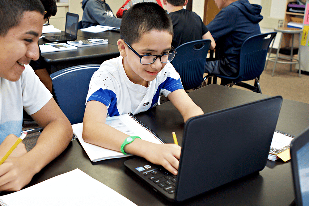Two students, one using a laptop and the other smiling at his screen, are engaged in a tutoring session with Amplify Tutoring at a table in a classroom setting.