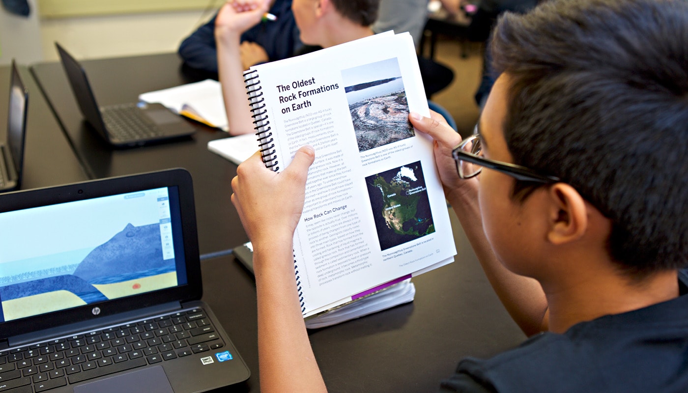 A student holding a textbook about rock formations while using a laptop in a science classroom.