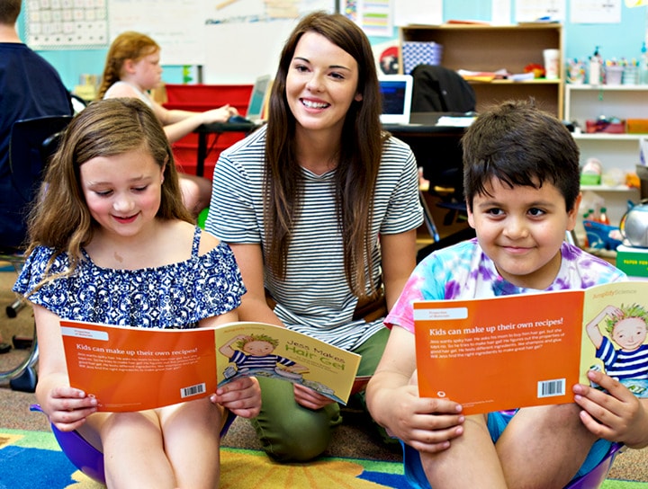 Teacher sitting on the floor with two students, all smiling and reading colorful books in a science classroom.