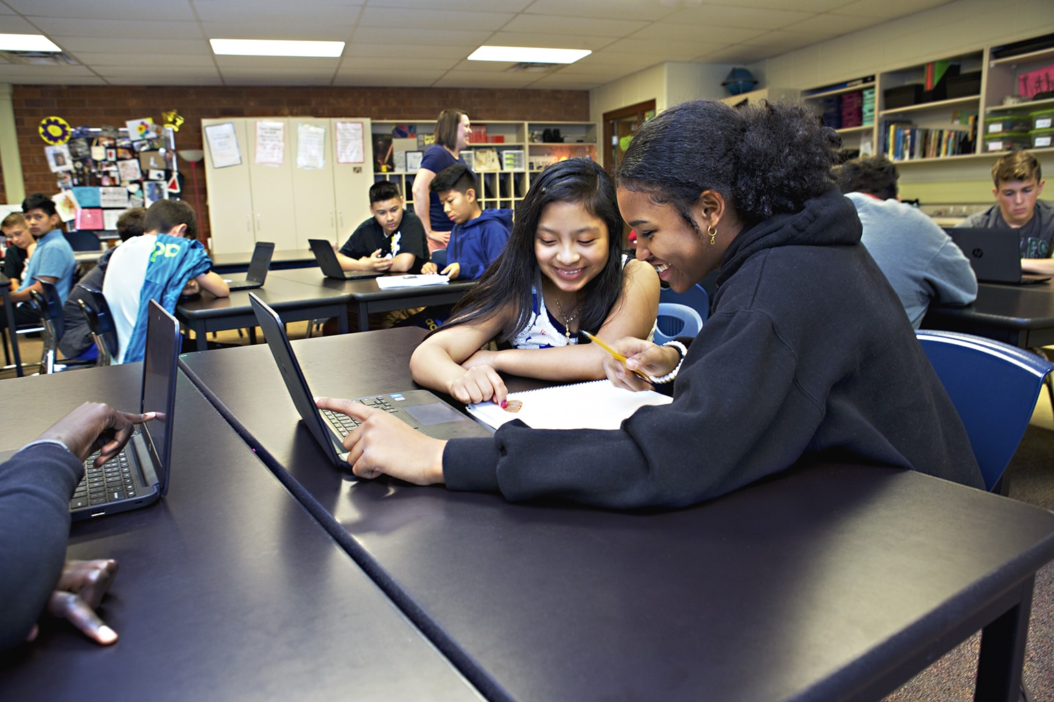 Two students smiling and working together at a desk in a classroom with other students and laptops around them.