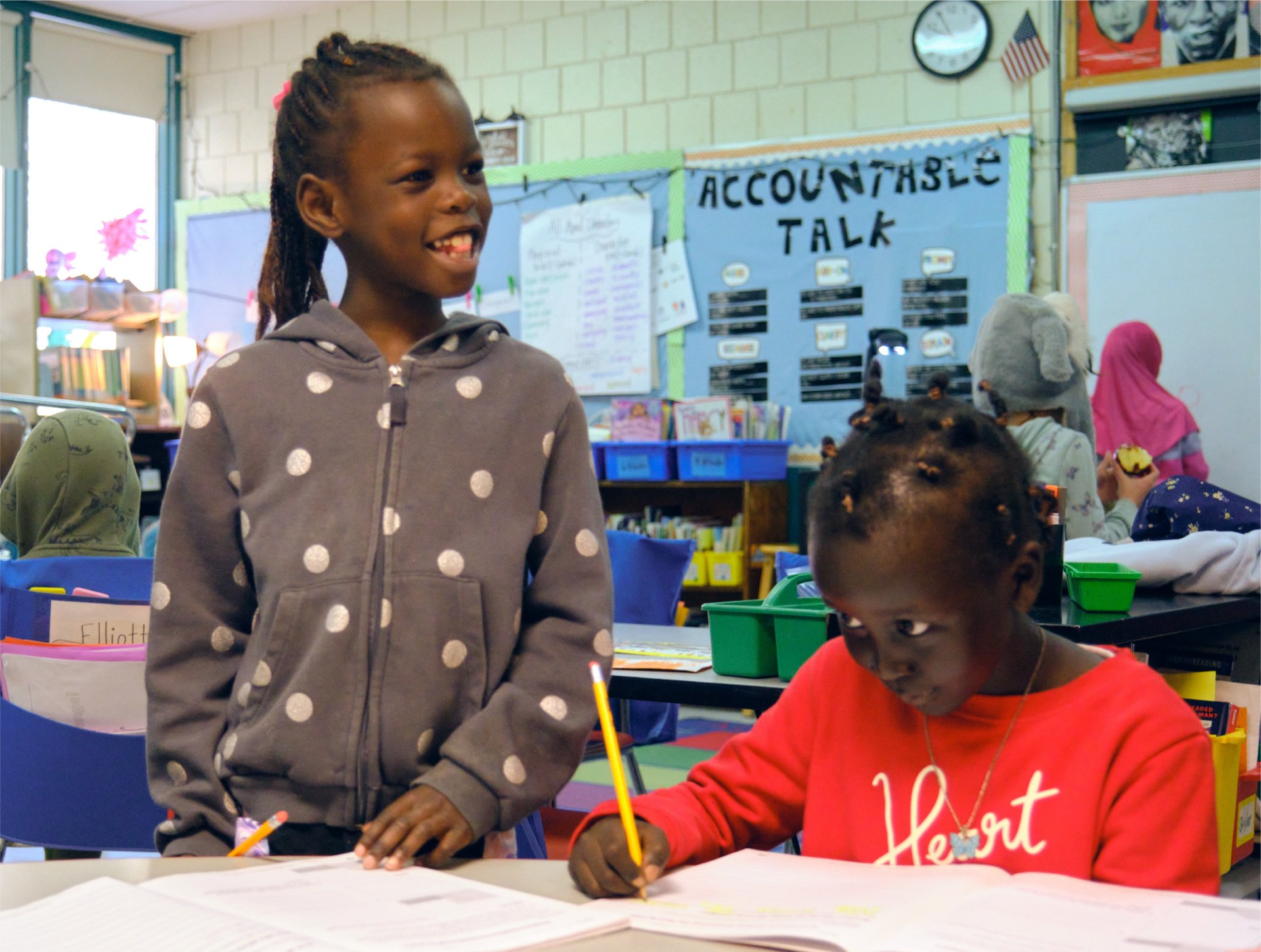 Two young girls in a literacy intervention classroom setting; one standing and smiling, and the other seated, focused on writing in a notebook.