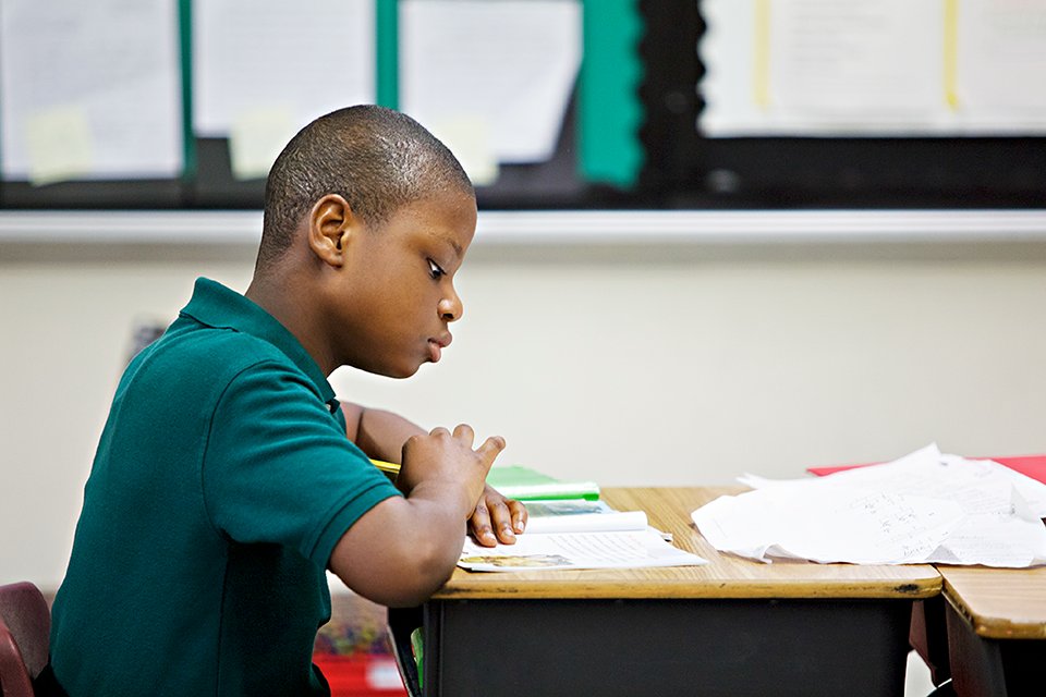 A young boy in a green shirt concentrates on decoding a book at his school desk, surrounded by scattered papers.
