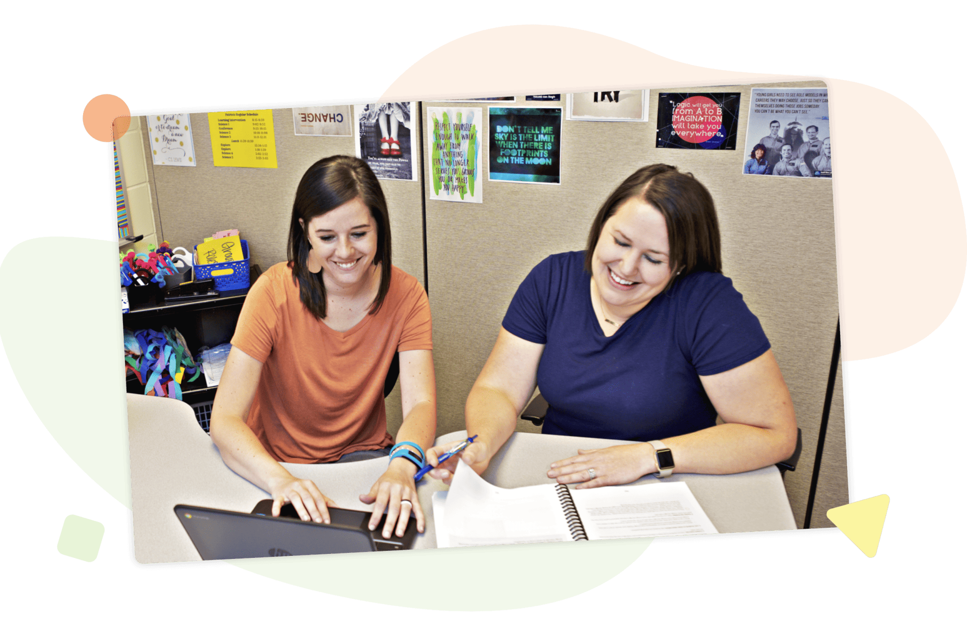 Two math teachers smiling and working together at a desk with a laptop and notebook, surrounded by colorful decorations and posters.