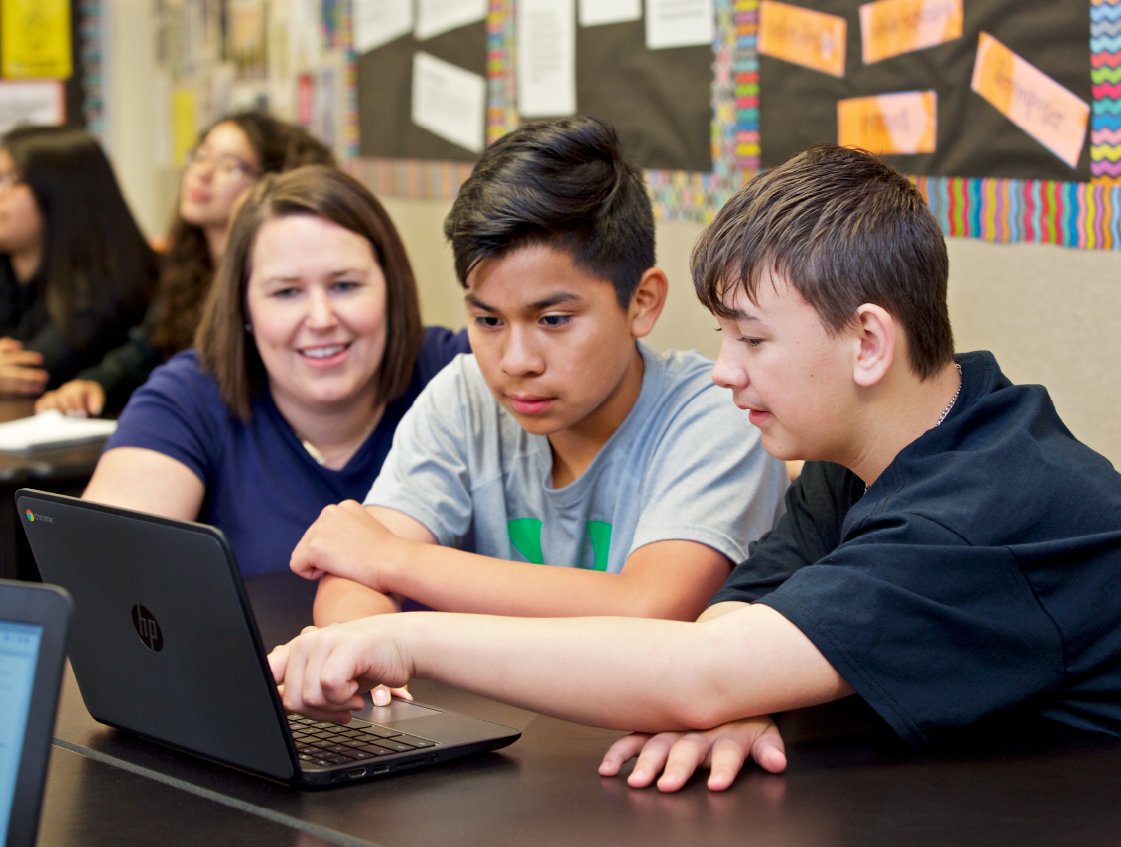 Two teenage boys and a female teacher using a laptop together in a classroom, with educational posters focused on the science of reading curriculum in the background.