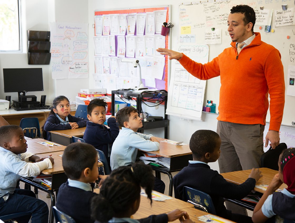 A teacher gesturing while explaining a lesson from a science of reading curriculum to attentive elementary students in a classroom filled with educational posters.