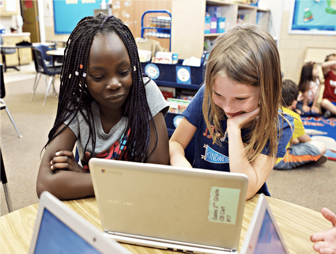 Two young girls, one with braided hair, using a laptop together in a classroom focused on early literacy with other students in the background.