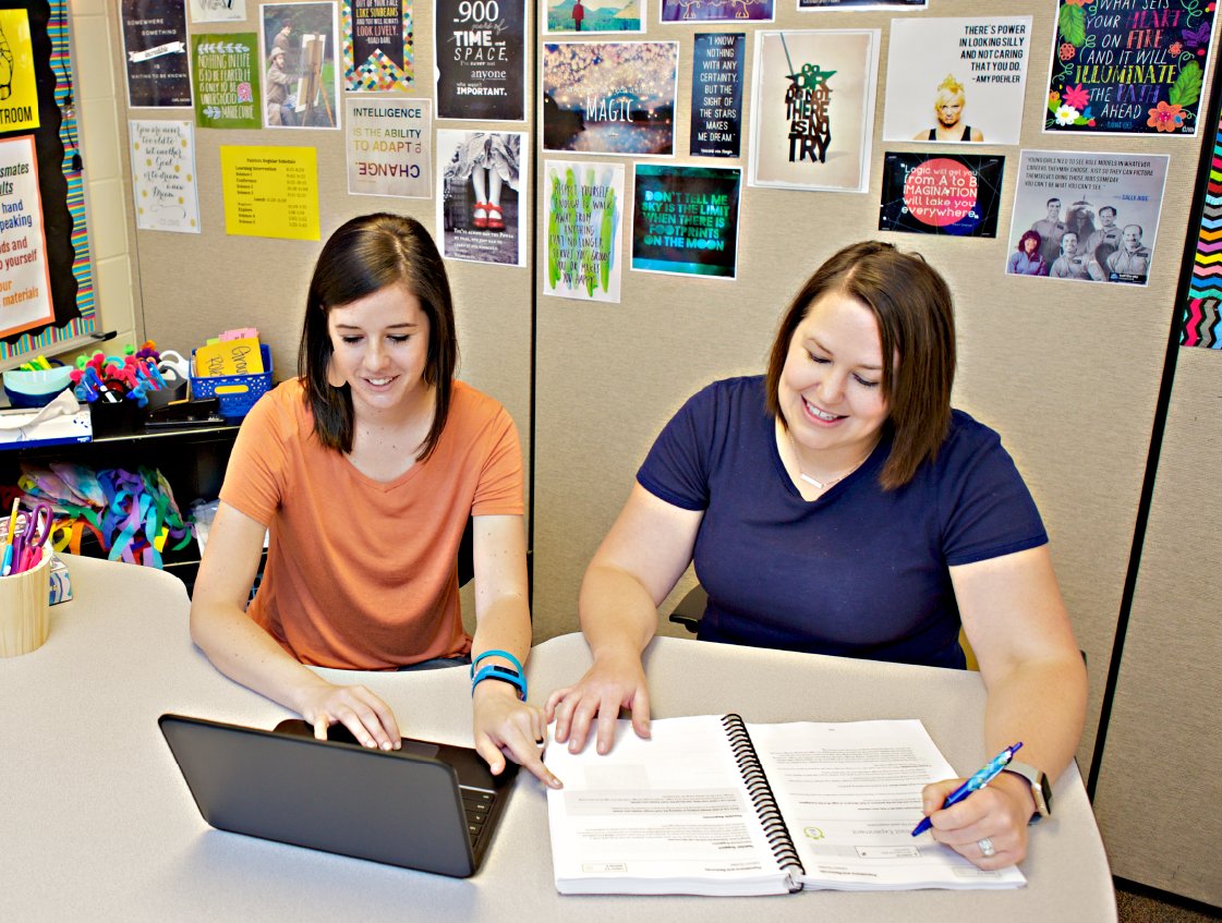 Two women smiling and working together at a desk with a laptop and notebook in an office decorated with posters about the science of reading curriculum.