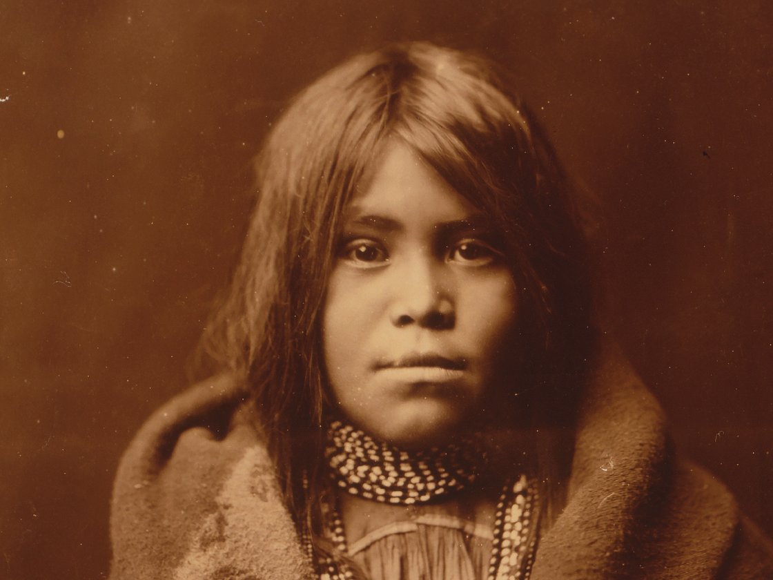 Vintage sepia-toned portrait of a young indigenous girl wearing a beaded necklace and wrapped in a blanket.