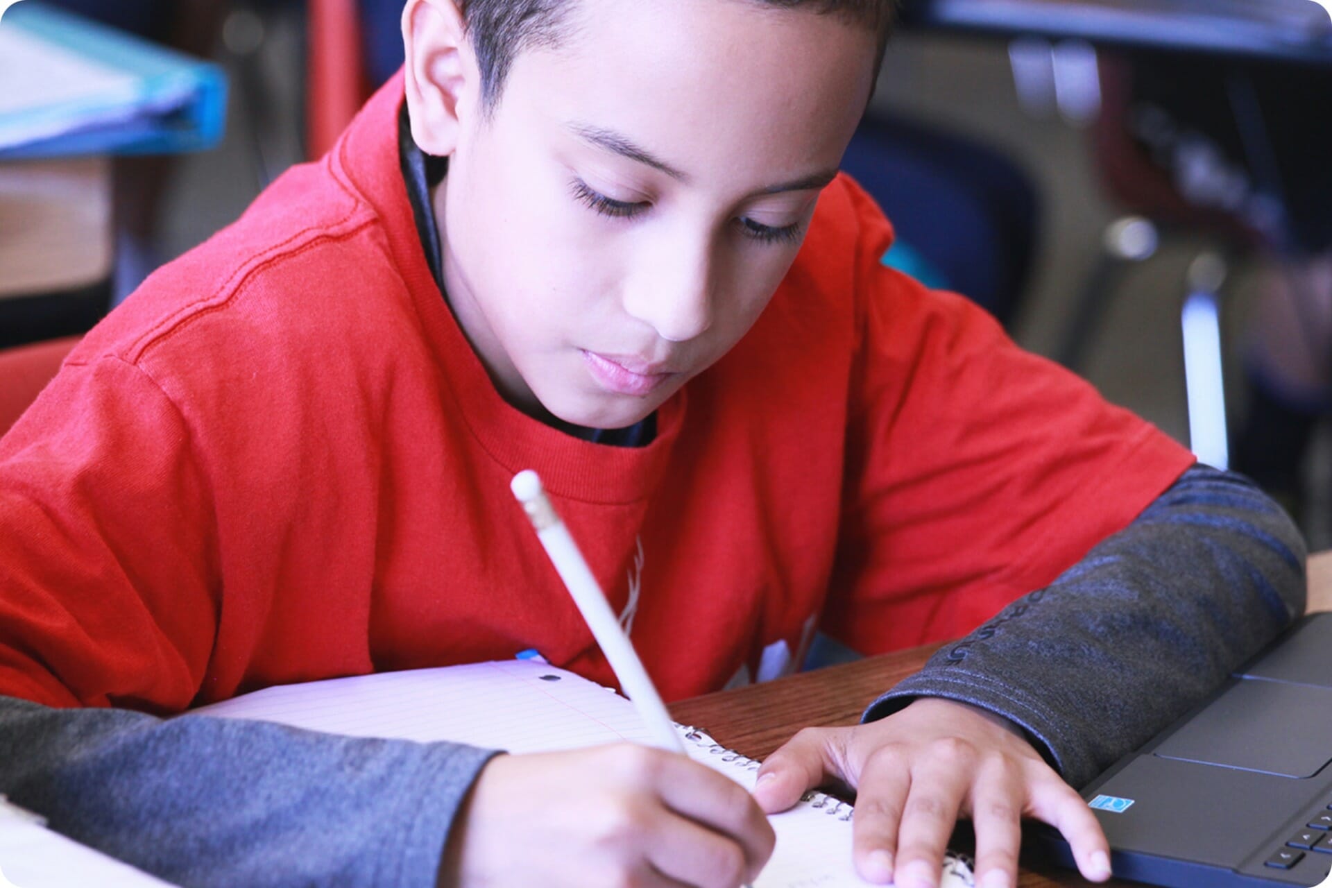 A young boy in a red hoodie writes in a notebook while using a laptop in a math classroom setting.