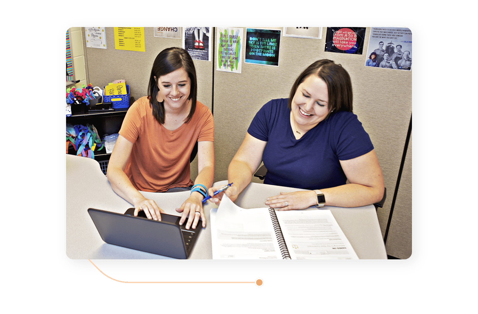 Two women collaborating in an office cubicle, using a laptop and reviewing documents for a reading fluency assessment.