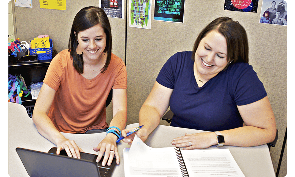 Two women in an office setting, smiling and working together with High Quality Instructional Materials, one using a laptop and the other writing in a notebook.