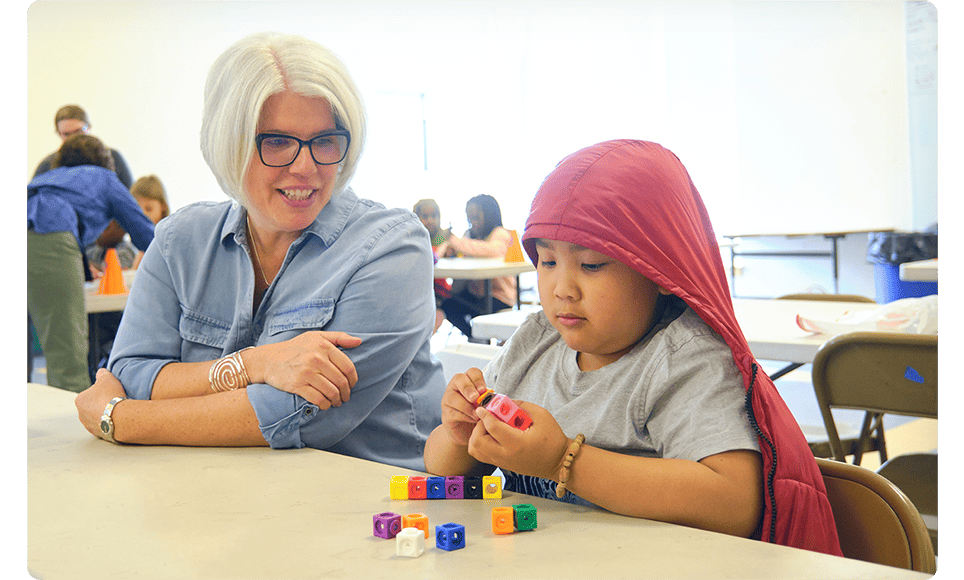 A woman in a blue shirt and glasses smiles while observing a young girl in a red headscarf playing with colorful blocks at a classroom table with High Quality Instructional Materials.