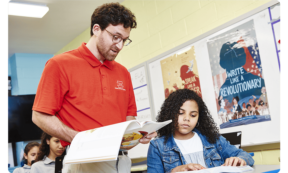 A teacher in a red shirt and glasses reading a book to a young girl in a classroom, with High Quality Instructional Materials on the wall behind them.