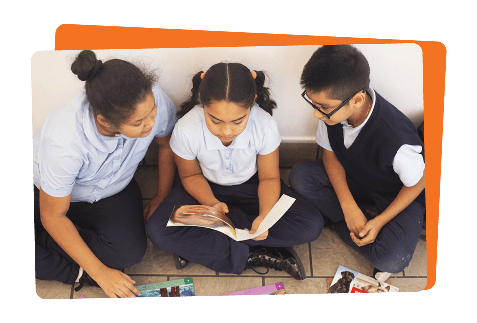 Three students in school uniforms sitting on the floor, reading a book together.