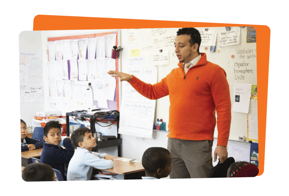 A teacher in an orange sweater presenting in a classroom, pointing to educational posters, with students attentively listening.