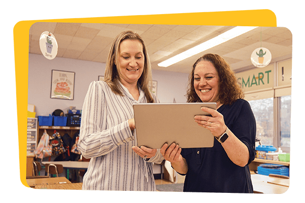 Two women in a classroom involved in professional development for teachers, looking at a tablet and smiling. One is holding the tablet. Classroom elements like posters are visible in the background.
