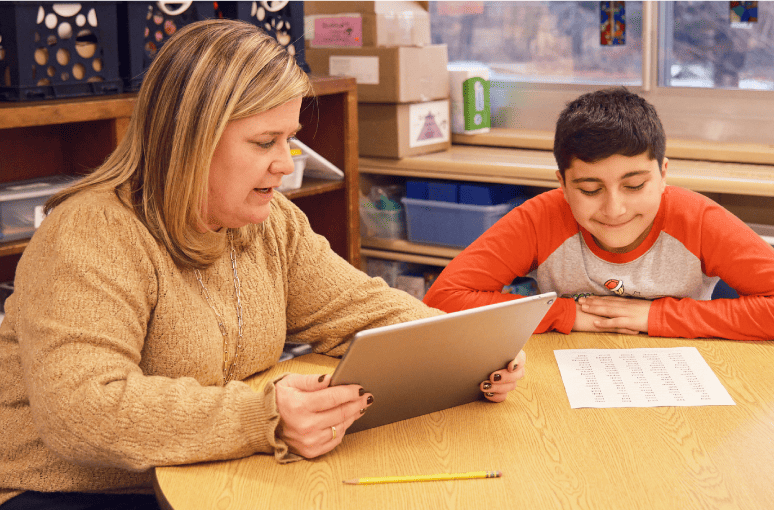 A woman and a young boy sit at a desk in a classroom, looking at a tablet together as part of the Back to School program, with papers and pencils on the desk.