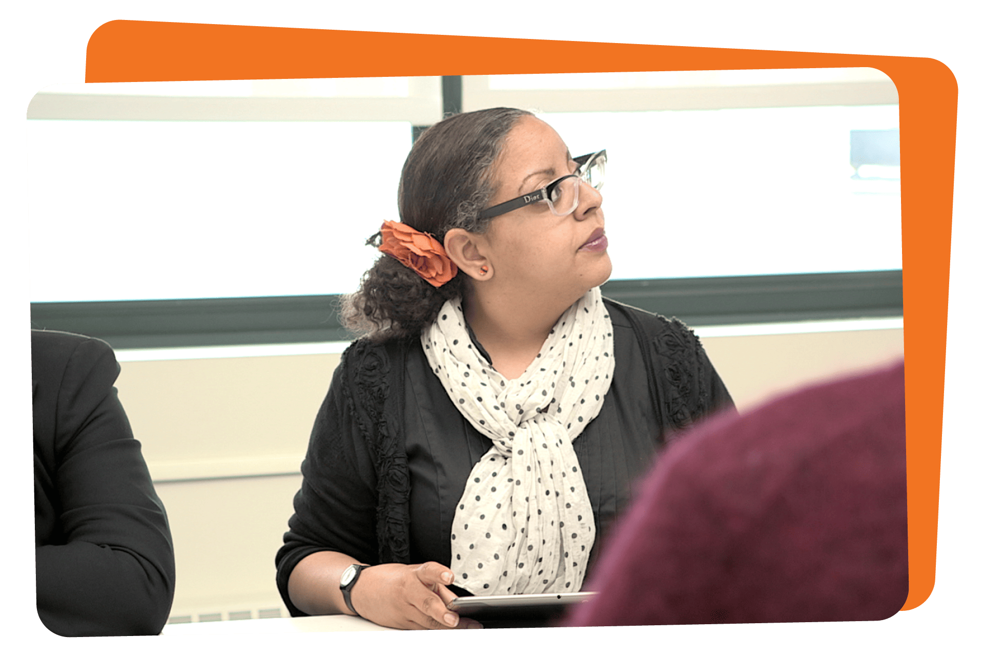 Woman with glasses, wearing a scarf and a flower in her hair, attentively listening in a classroom setting.