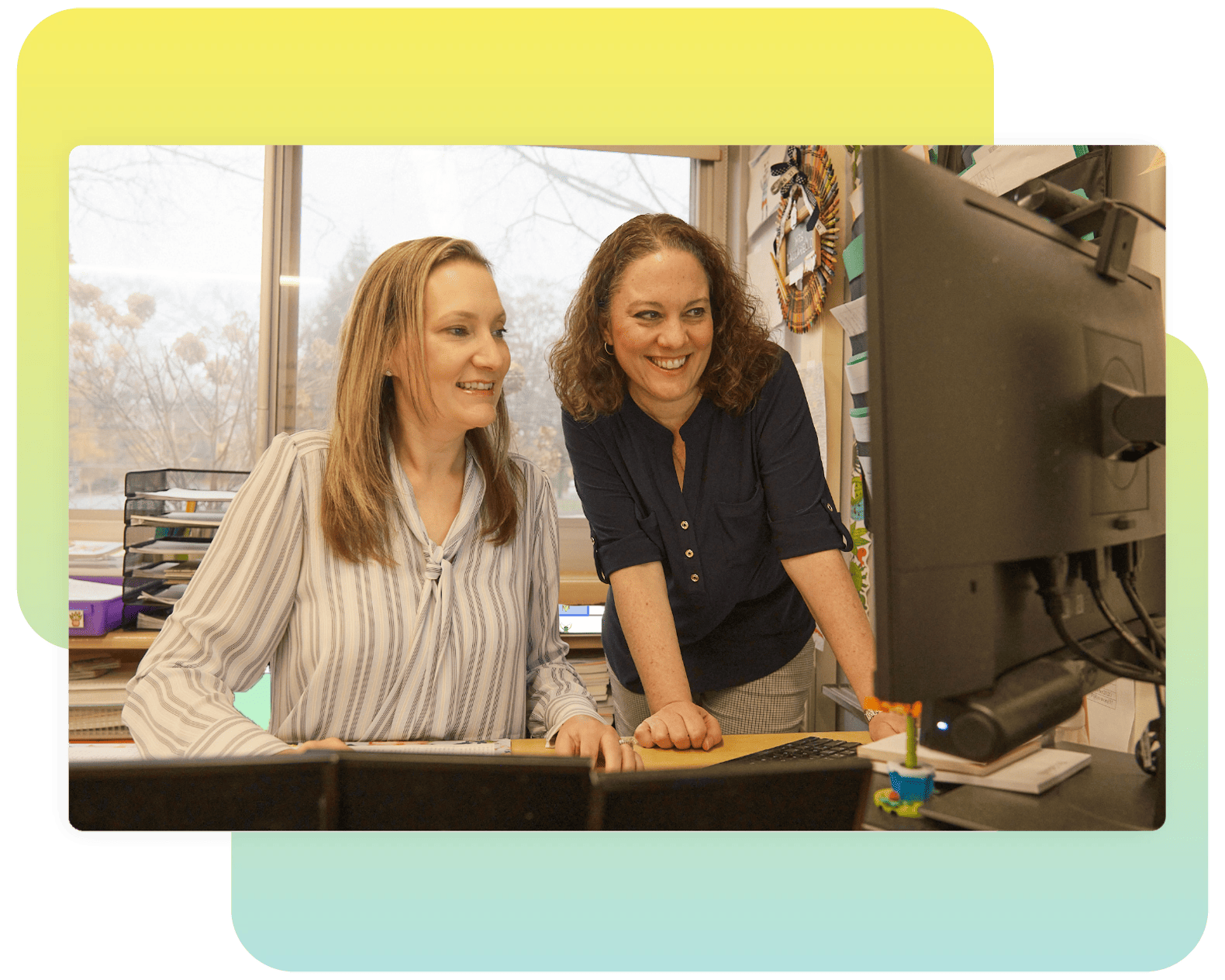 Two women smiling while working together at a computer on New York math in a bright office setting.