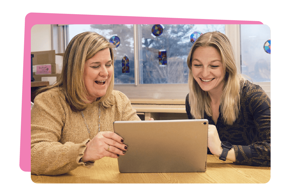 Two women smiling and looking at a tablet together in an office setting with colorful window decorations in the background, exploring Amplify change management curriculum resources.