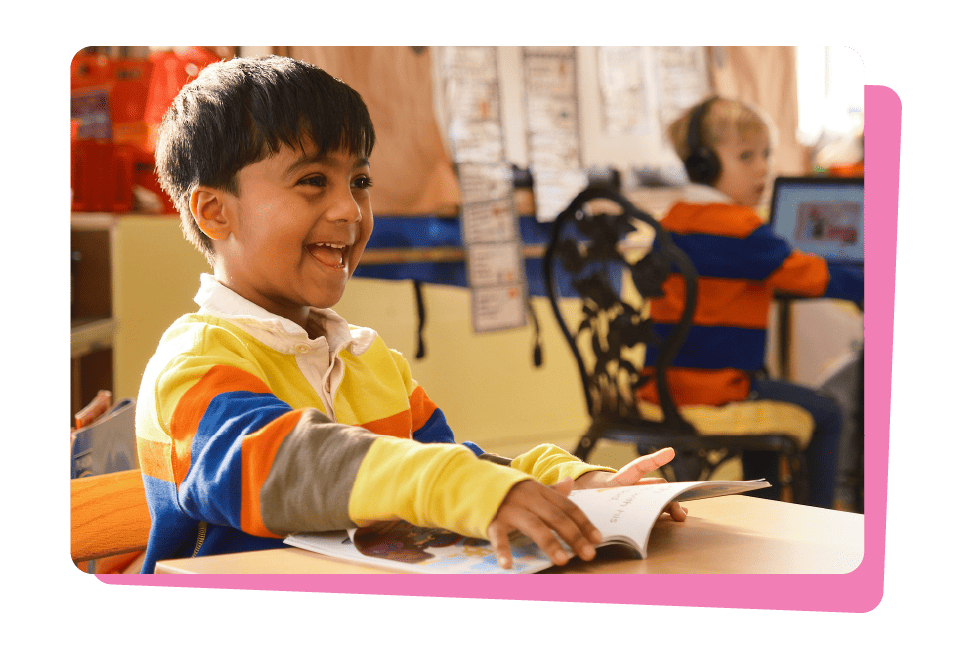 A young boy in a colorful striped sweater smiles while reading a book from the Amplify curriculum at a classroom desk, another child in the background, representing science of reading success stories.