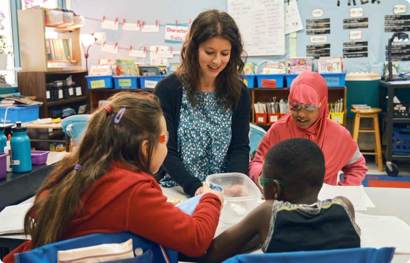 In a vibrant classroom setting, a teacher engages with three K—5 students in a group activity at a table. Elements like bookshelves and posters enrich the scene, while Amplify Science materials seamlessly integrate into their hands-on experience.