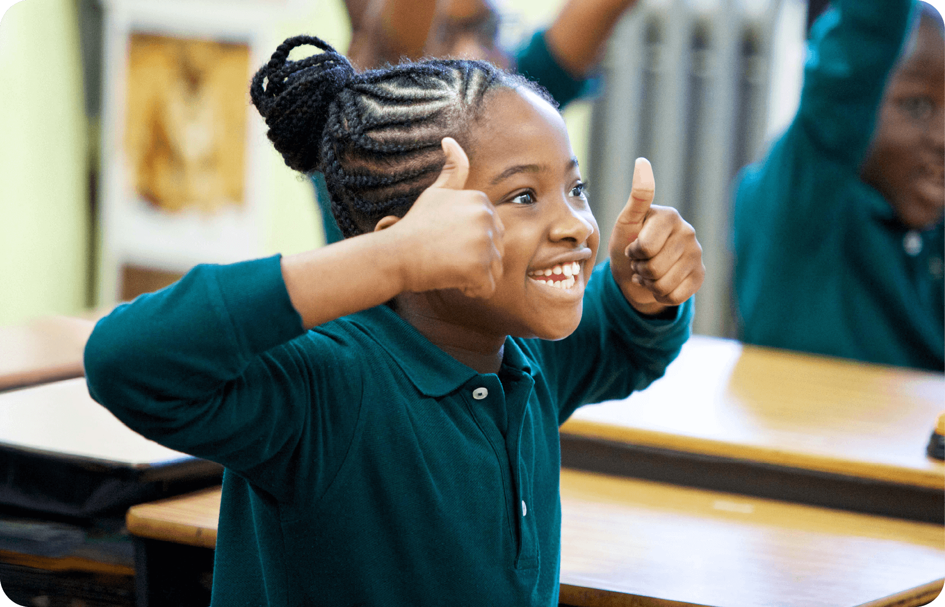 A child in a green shirt smiles and shows two thumbs up while sitting at a desk in a classroom, enjoying the engaging atmosphere of the catholic school curriculum.